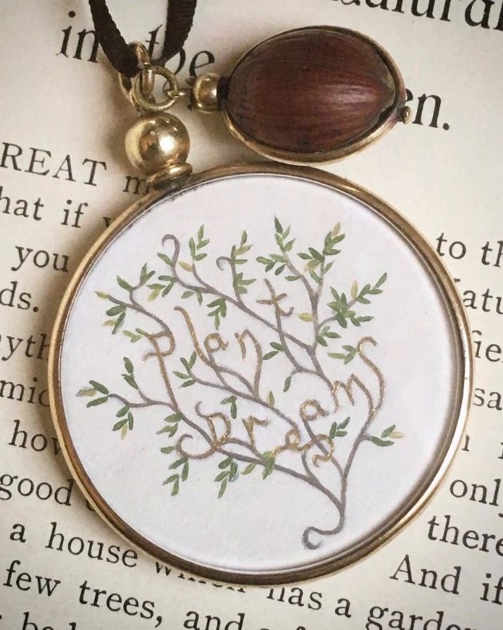 An Edwardian double locket painted with branches spelling Plant dreams, hung beside a wooden acorn charm on an open book