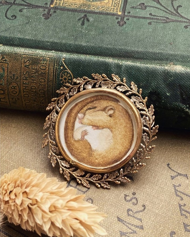 A painted hazel dormouse curled asleep inside an ornate leafy Edwardian locket brooch, resting against a green cloth-bound book
